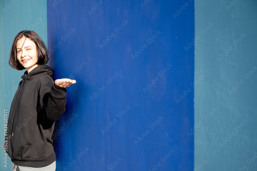 Modern girl in black sweatshirt isolated on plain bright blue background, pointing with hand to side empty space for table or chart. Photo of model on wall background with empty space for text.