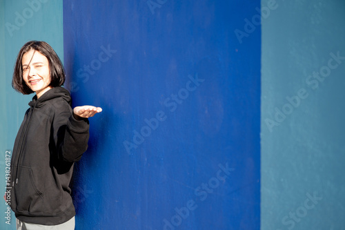 Obraz na płótnie Modern girl in black sweatshirt isolated on plain bright blue background, pointing with hand to side empty space for table or chart. Photo of model on wall background with empty space for text. w salonie