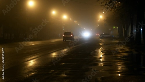 Car traffic in fog on a dark wet gloomy city road with poor visibility at night illuminated by lanterns and headlights 