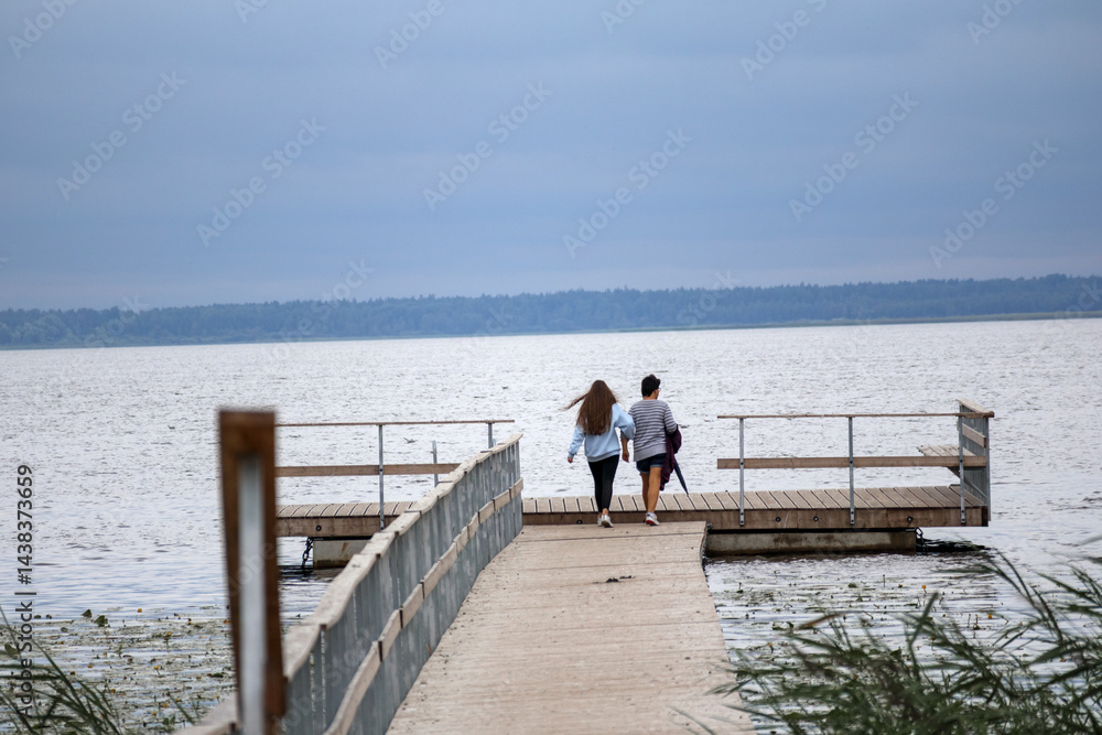 Two people, a man and a woman, walking from behind along a wooden pier towards a calm body of water under a cloudy sky.