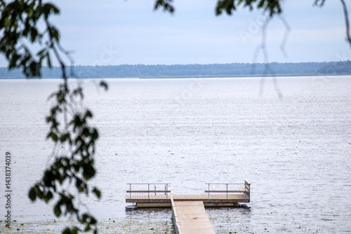 Obraz na płótnie
a wooden pier stretching into calm water, with a small platform at the end and blurred tree branches in the foreground under a cloudy sky. w salonie