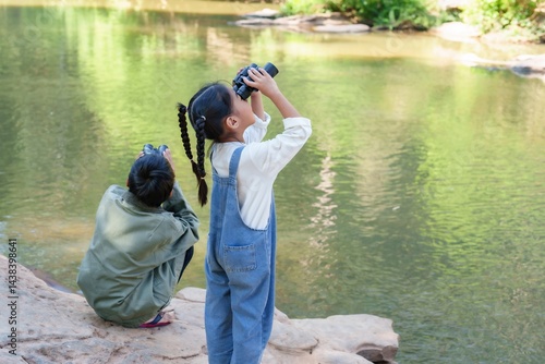 Asian young girl and Asian boy using binoculars for birdwatching activity near riverbank in forest, expressing joy, outdoor adventure, exploration spirit, bonding with nature, environmental awareness.