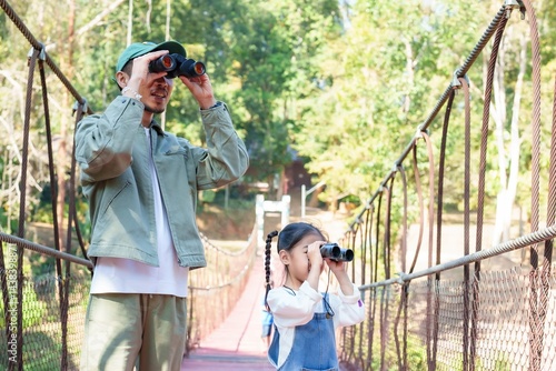Asian adult man and young Asian girl standing side by side using binoculars for birdwatching on suspension bridge in forest, expressing excitement, connection, outdoor learning, nature exploration,