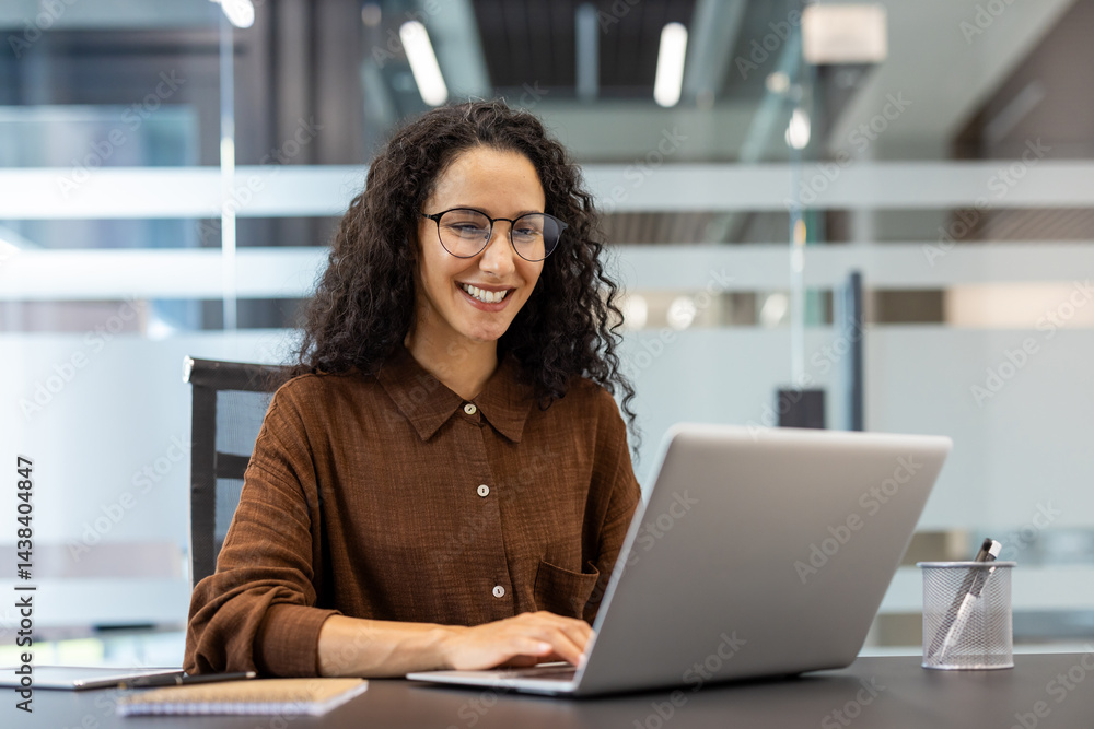 A cheerful woman with glasses works at a desk in a modern office, focused on her laptop with a big smile. The office is bright