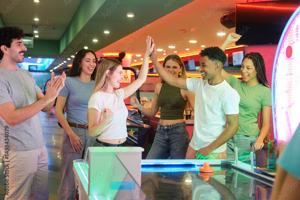 Group of friends giving high five while playing air hockey, celebrating victory in amusement park