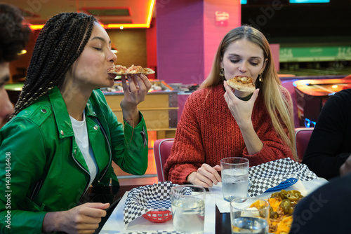 Obraz na płótnie Two young women enjoying pizza slices at a bowling alley restaurant, sharing a meal and good times w salonie
