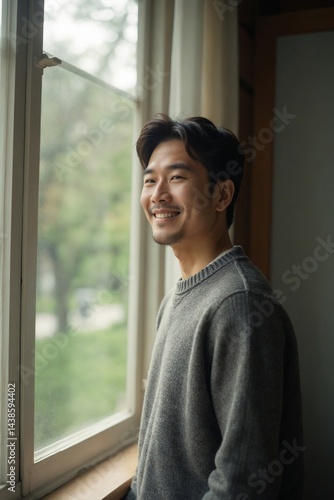 Free Download Smiling Asian Man in Casual Sweater Looks Out Large Window Towards Blurry Park View, Relaxed Home Lifestyle - GFXLoad