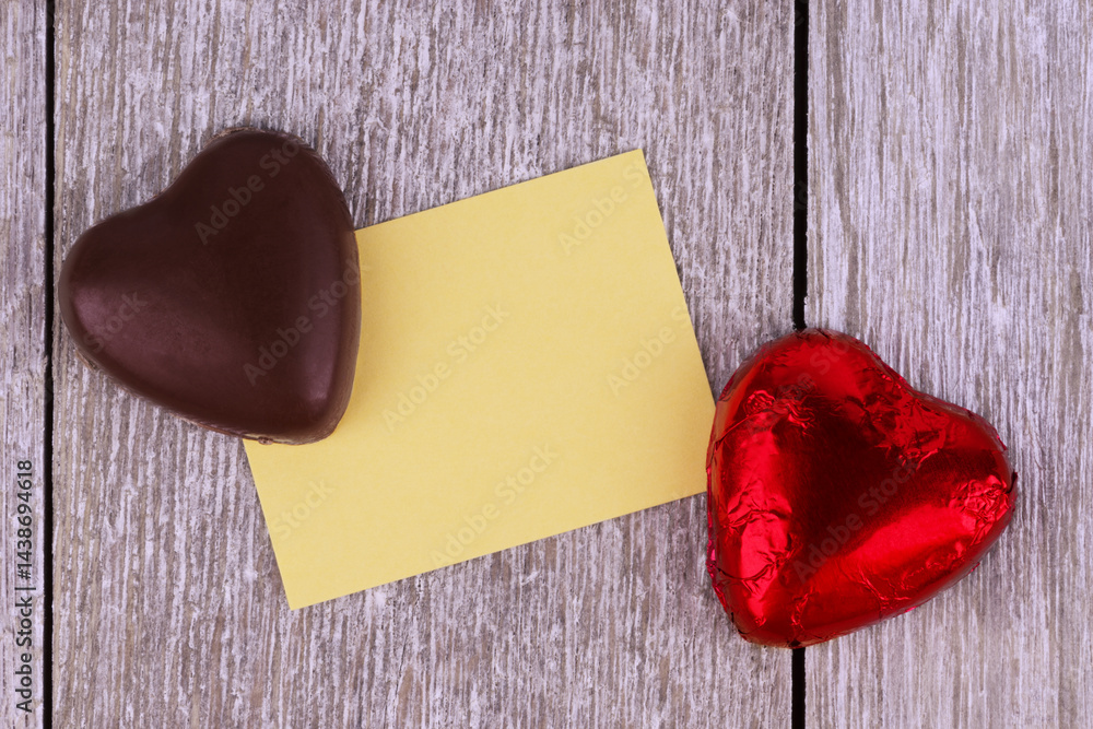 Two heart-shaped chocolates, one wrapped in red foil, placed on a rustic wooden background with a blank yellow card, perfect for Valentine's Day, love messages, or romantic celebrations.