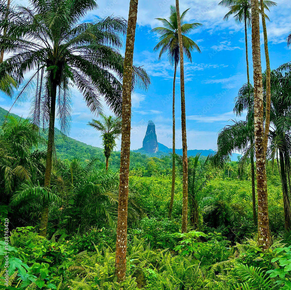 Obraz premium Tropical jungle view of Pico Cão Grande, a dramatic volcanic spire rising from lush greenery under a bright blue sky in São Tomé and Príncipe. Exotic, untouched natural paradise.
