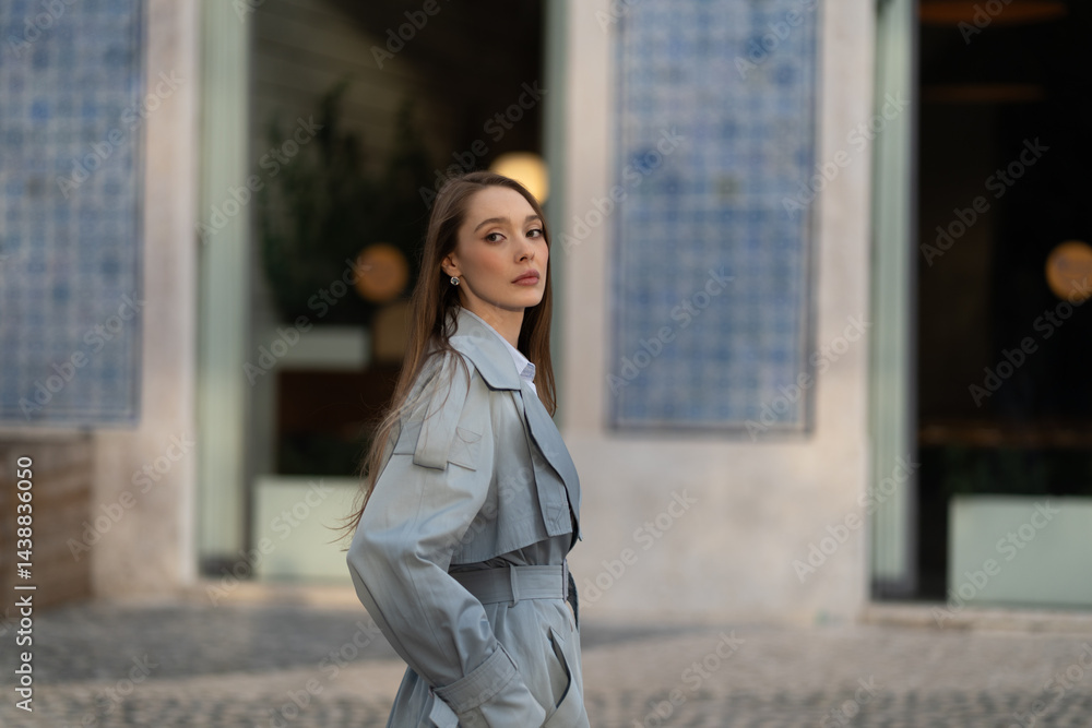 Fototapeta premium Side view of stylish young woman in blue outfit looking at camera. Beautiful lady with long hair walking against blurred buildings in city