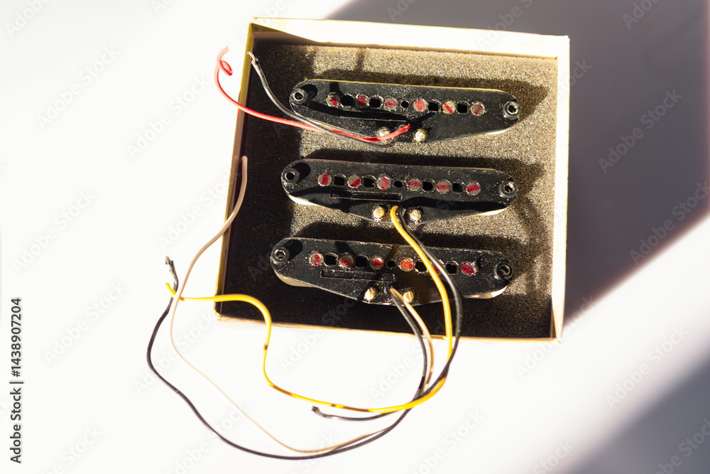 Three black single-coil guitar pickups with red pole pieces sit in a foam-lined box, casting shadows on surface. Wires extend from each pickup, ready for wiring into an electric guitar