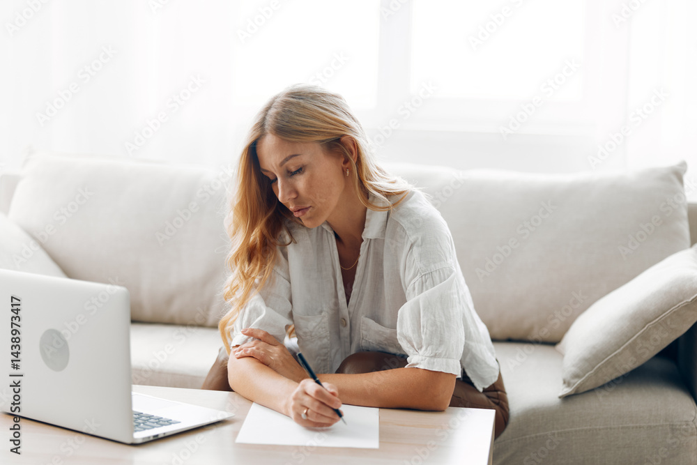 Woman working from home, writing notes on paper while sitting on a comfortable couch with a laptop nearby Woman working from home, writing notes on paper while sitting on a comfortable couch with a laptop nearby