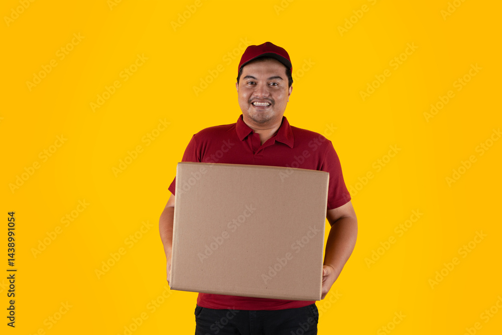 Smiling delivery man wearing red uniform and cap with delivery box on yellow background Smiling delivery man wearing red uniform and cap with delivery box on yellow background