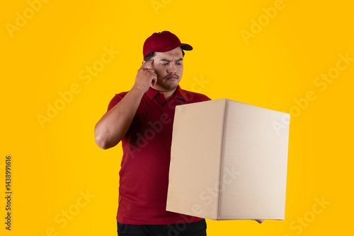 Confused delivery man wearing red uniform and cap with delivery box on yellow background