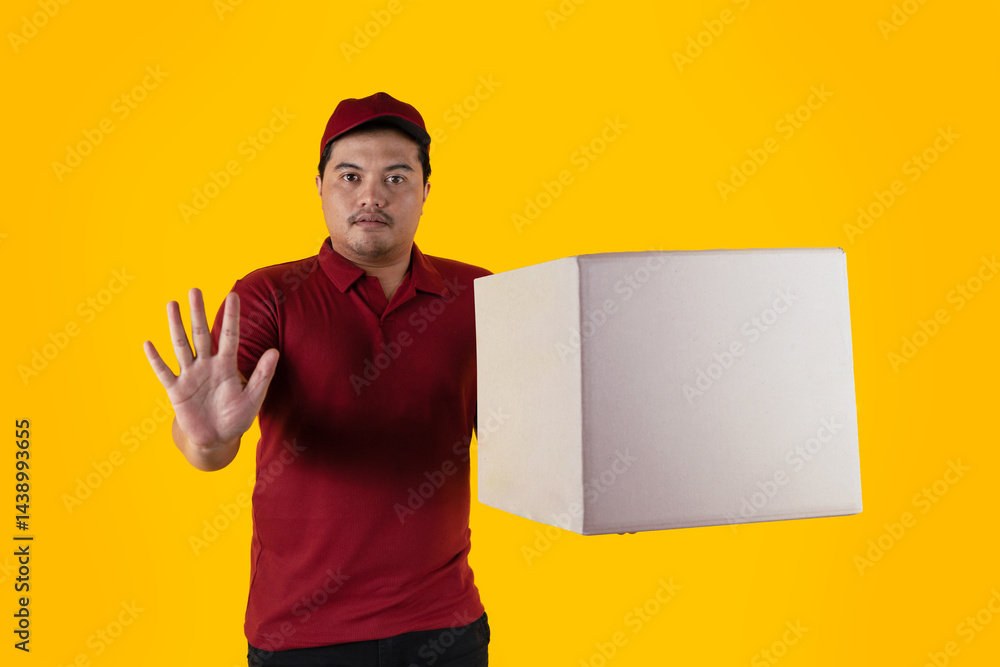 Delivery man with stop hand gesture wearing red uniform and cap with delivery box on yellow background Delivery man with stop hand gesture wearing red uniform and cap with delivery box on yellow background