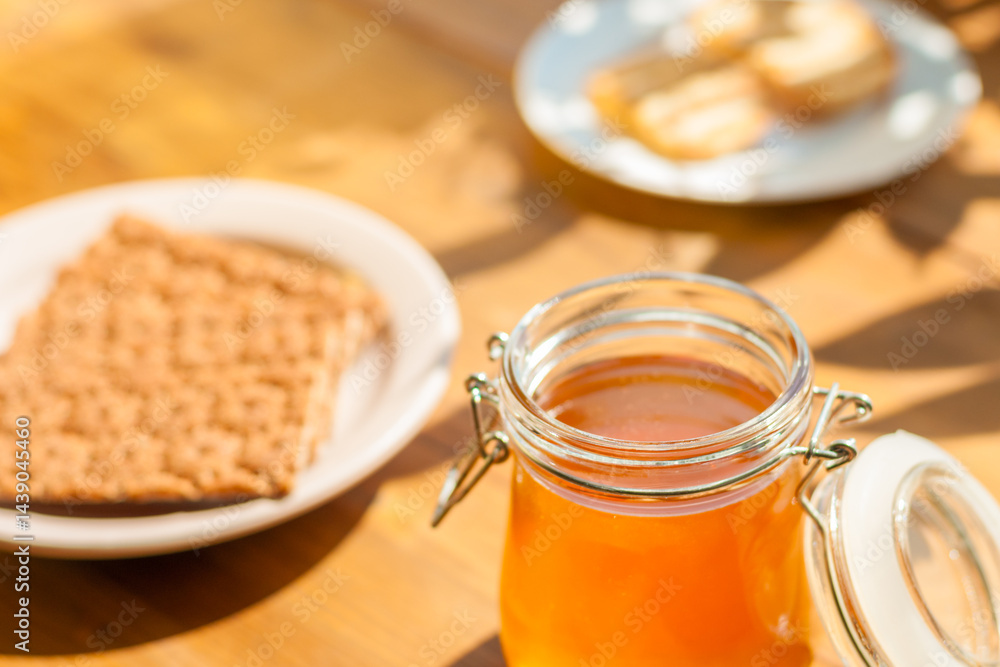 A glass jar filled with golden honey rests on a wooden table, while a plate of crackers and cookies compliments the inviting sunlight ambiance.