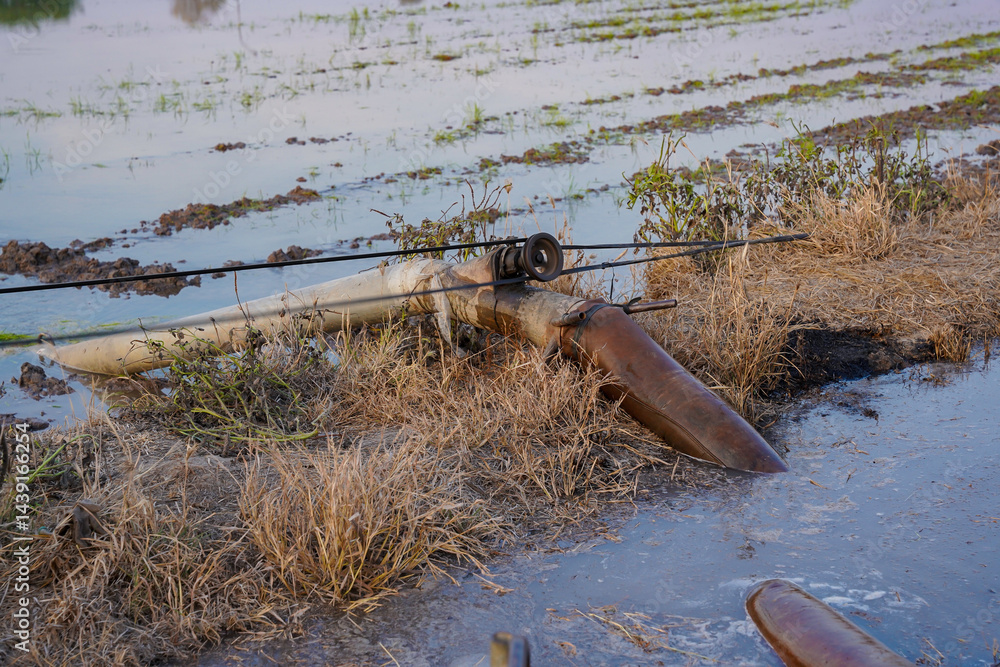 Fishing kayak with rod and paddle resting on muddy riverbank, surrounded by dry grass and water, peaceful outdoor scene