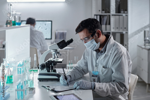 Scientist wearing protective gear and a face mask, recording observations in lab. Various scientific equipment and colorful chemical samples visible in workspace