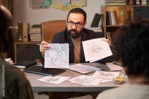 Bearded school psychologist showing child drawings with aggressive symbols to father of schoolboy at therapy session