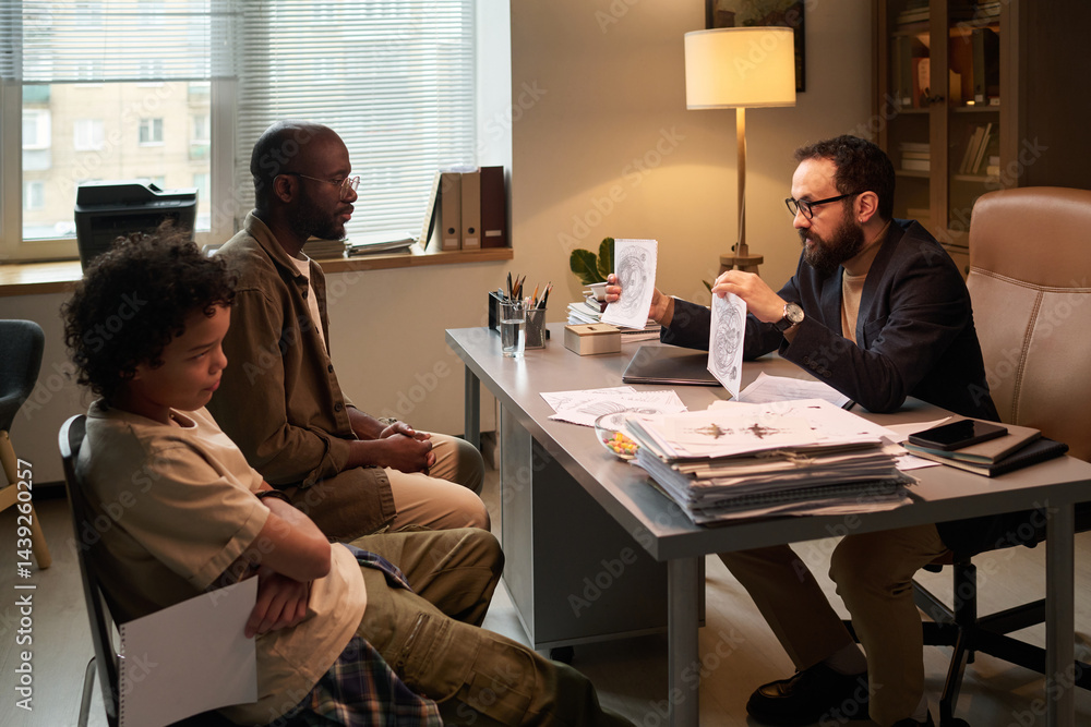 African American man and his son looking at child drawings held by mature school psychologist during session