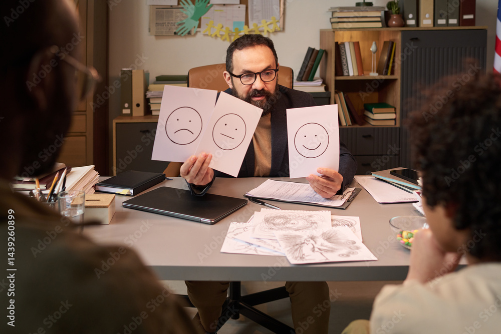 Confident psychologist talking to schoolchild and asking him choose one of three different emotions illustrated on papers
