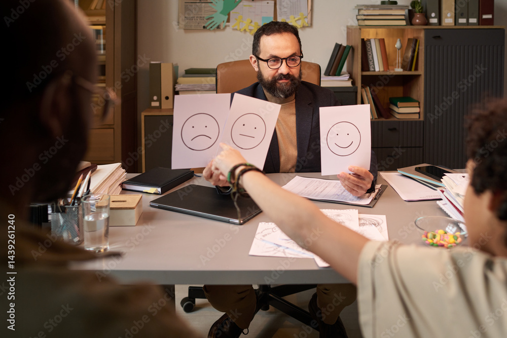Confident male psychologist holding three papers with various emotions and looking at schoolboy during therapy session