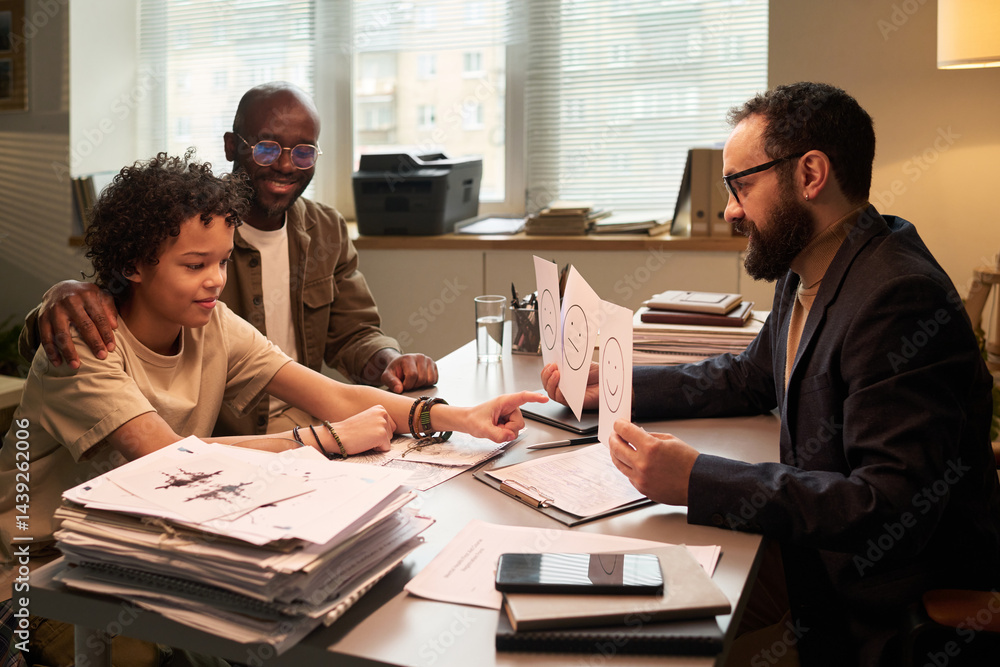Cute schoolboy pointing at paper with smiling face while sitting next to his father during communication with psychologist