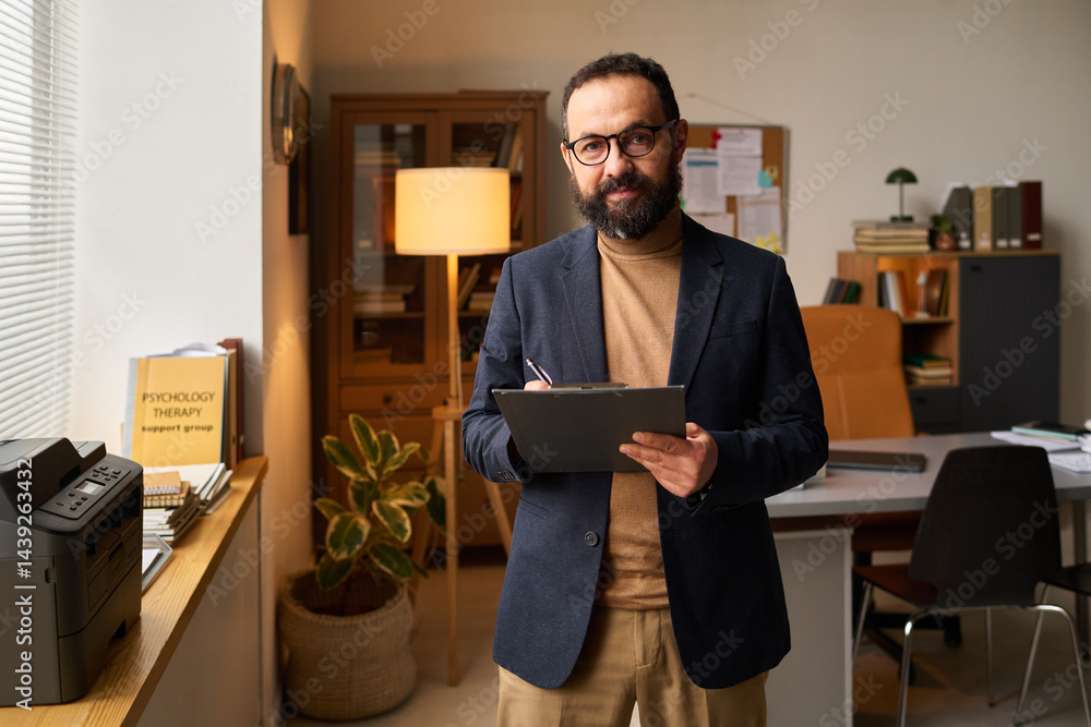 Successful male psychologist in smart casual attire taking notes in document and looking at camera against his workplace