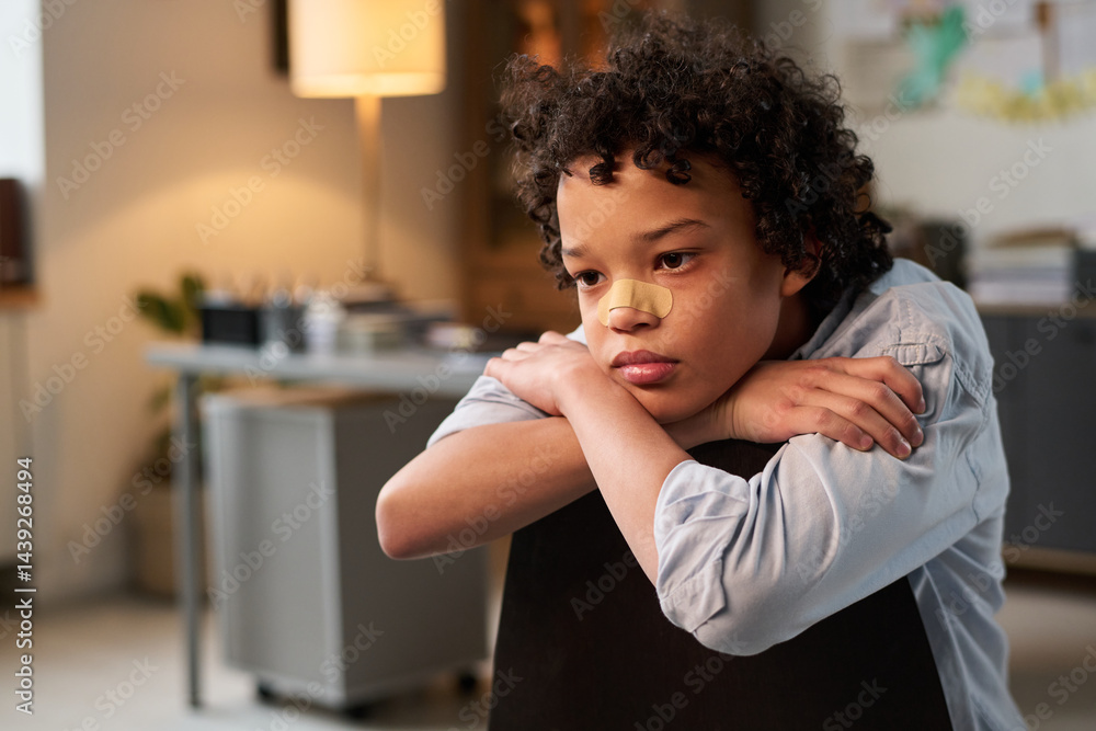 Sulky African American boy with bandage on nose sitting on chair in psychologist office and thinking of his behavior
