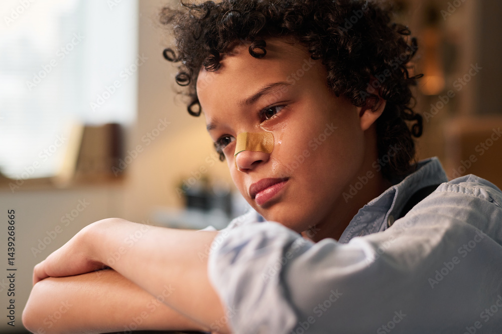 Miserable schoolboy with bandage on his nose sitting in front of camera and crying while visiting school psychologist