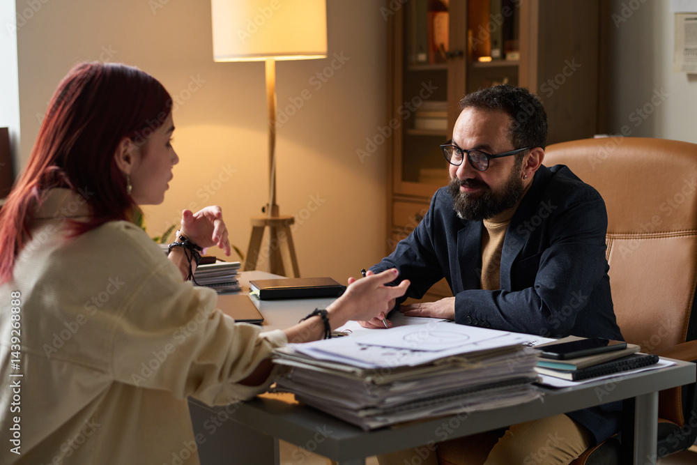 Smiling bearded male psychologist looking at schoolgirl sitting in front of him and describing her problem at session in office