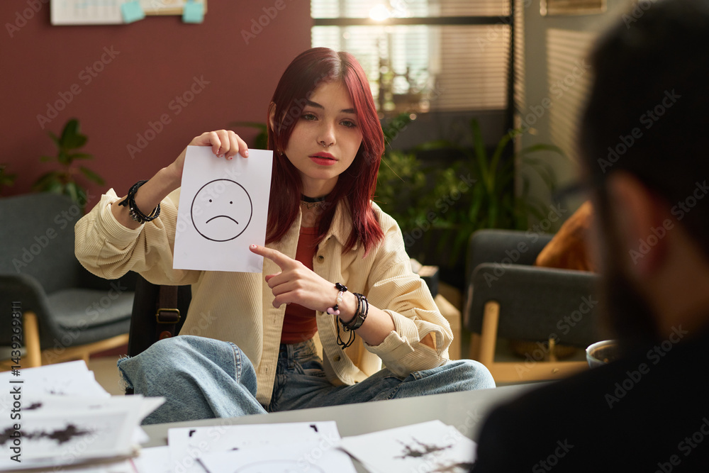 Teenage girl pointing at paper with sad smile and looking at school psychologist while explaining him her emotions