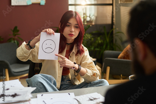 Obraz na płótnie Teenage girl pointing at paper with sad smile and looking at school psychologist while explaining him her emotions w salonie