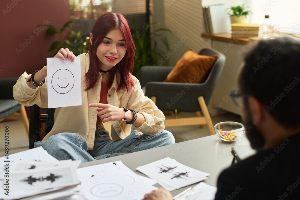 Happy teenage girl pointing at paper with smiling face and looking at school psychologist during psychological session