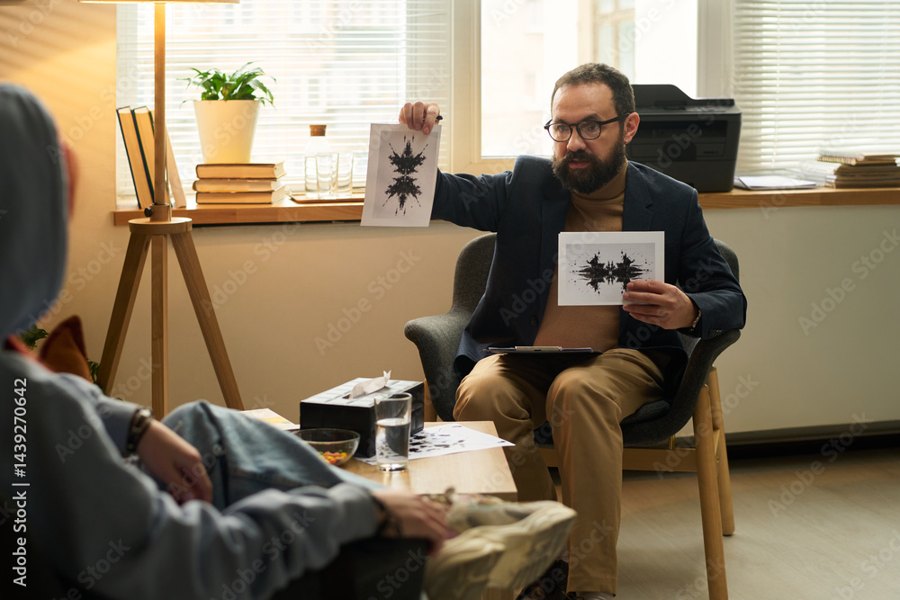 Bearded psychologist in eyeglasses showing papers with inkblots to teenage patient during psychological session in office