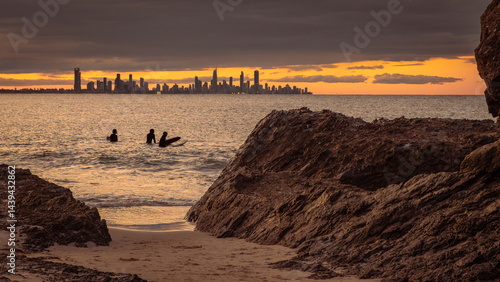Three framed surfers in silhouette wait for a wave at Currumbin alley with Surfers Paradise in the distance across the bay under threatening clouds as the sun sets on the Gold Coast, Australia.
