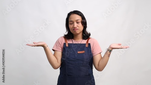 Young female barista wearing a navy apron making a confused expression, raising both hands in a shrug gesture. Studio shot with white background, representing doubt, uncertainty, or not knowing