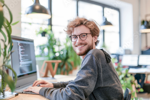 Smiling young man with glasses working on a computer in a bright, modern office filled with natural light and greenery