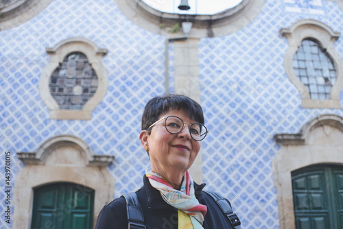Senior tourist wearing glasses and colorful scarf smiling in front of a traditional portuguese facade decorated with blue and white azulejos tiles, enjoying her travel adventure