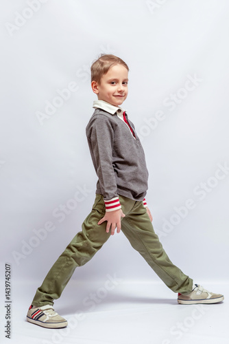 A child in a white-collared shirt and a gray sweater with red cuffs, standing in a dynamic pose, wearing green pants and sneakers, smiling confidently, facing slightly to the side