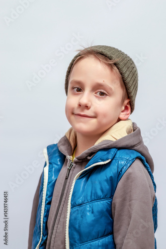 Smiling young child wearing a green beanie and blue puffy vest over a gray hoodie, looking at the camera with a neutral pale background. The child has short hair and a calm expression