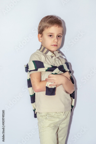 Young boy with short light brown hair wearing a beige shirt with a dark-striped sweater draped around shoulders, holds a piece of fabric, looking at camera, pale light background