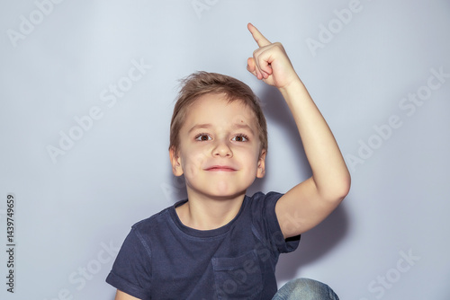 Young child with short hair looks upwards, smiling with a raised index finger. Outfit includes a dark short-sleeve shirt. Light backdrop creates a soft contrast with the child's expression