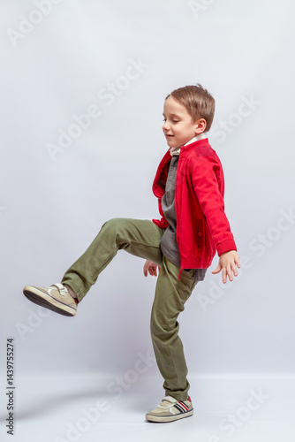 Young child lifts one leg in playful pose, wearing a bright red jacket, gray shirt, olive green pants, and beige sneakers with red stripes, against a neutral backdrop