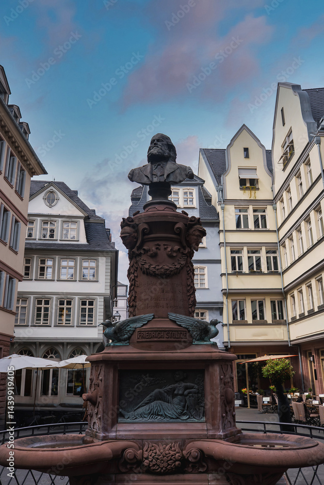 Statue of Friedrich Schiller atop an ornate fountain with intricate carvings, surrounded by European style buildings under a partly cloudy sky in Frankfurt.