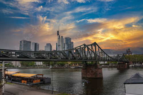 Obraz na płótnie Eiserner Steg spans the Main River in Frankfurt, Germany, with a docked boat, historic cathedral spire, and modern skyscrapers under an overcast sky. w salonie