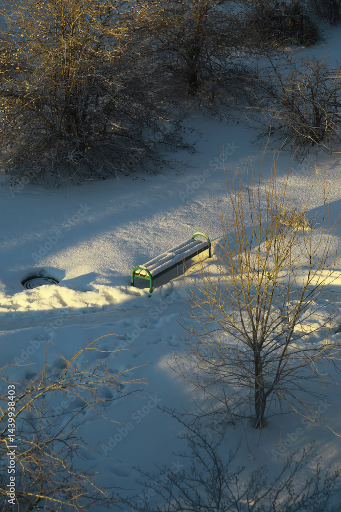 Winter Park Bench in Snowy Landscape with Footprints and Bare Trees at Sunset