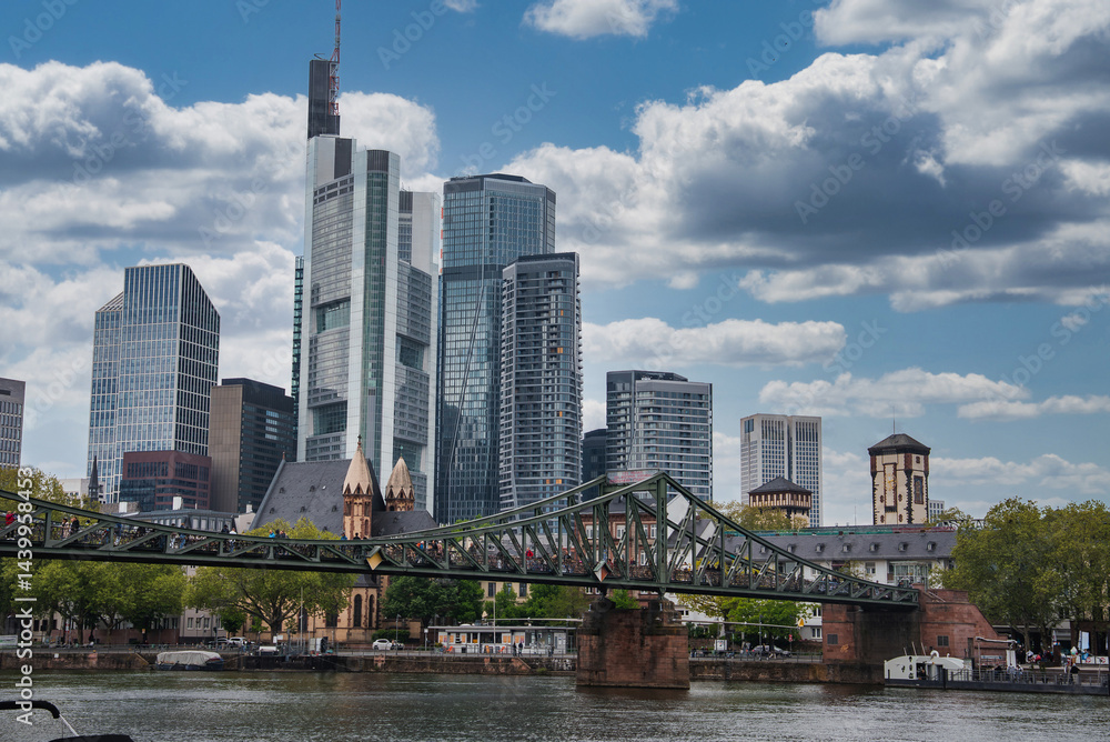 Skyline of Frankfurt, Germany, featuring Commerzbank Tower, modern skyscrapers, Eiserner Steg bridge over the Main River, and lush green trees.