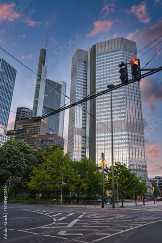 Frankfurt cityscape featuring Commerzbank Tower, reflective skyscrapers, a red traffic light, tram wires, green trees, and a cloudy sky.