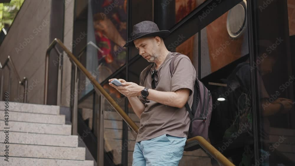 A middle-aged man in casual attire with a backpack stands on urban stairs, checking his smartphone for transport apps and maps. Captures modern city navigation A middle-aged man in casual attire with a backpack stands on urban stairs, checking his smartphone for transport apps and maps. Captures modern city navigation